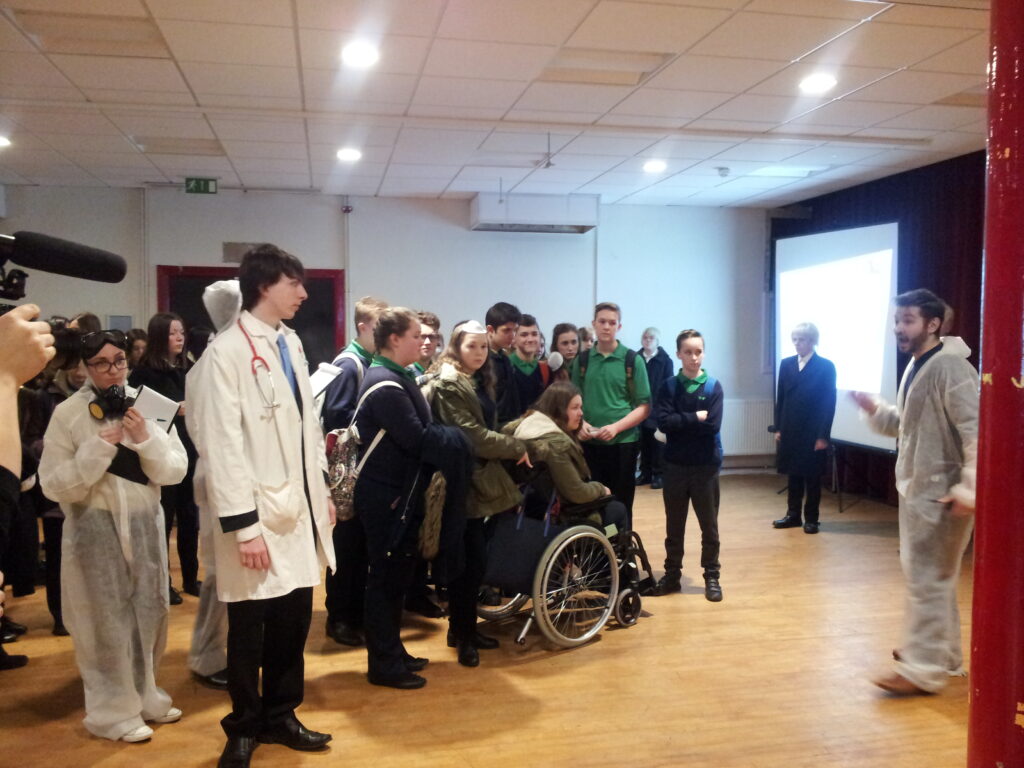 Pupils in a hall watching a presenter, some dressed in lab coats and protective suits